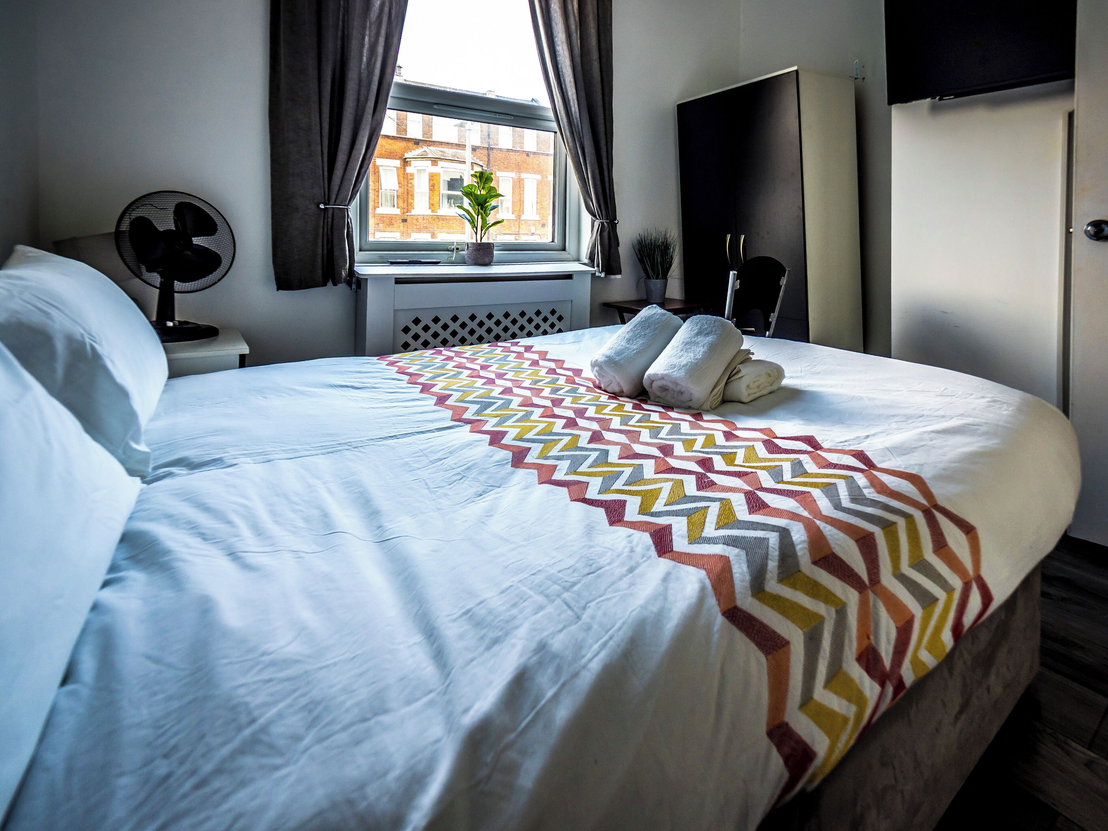 Una cama bien hecha con una colcha estampada, una ventana con cortinas, un ventilador y vistas a un edificio.