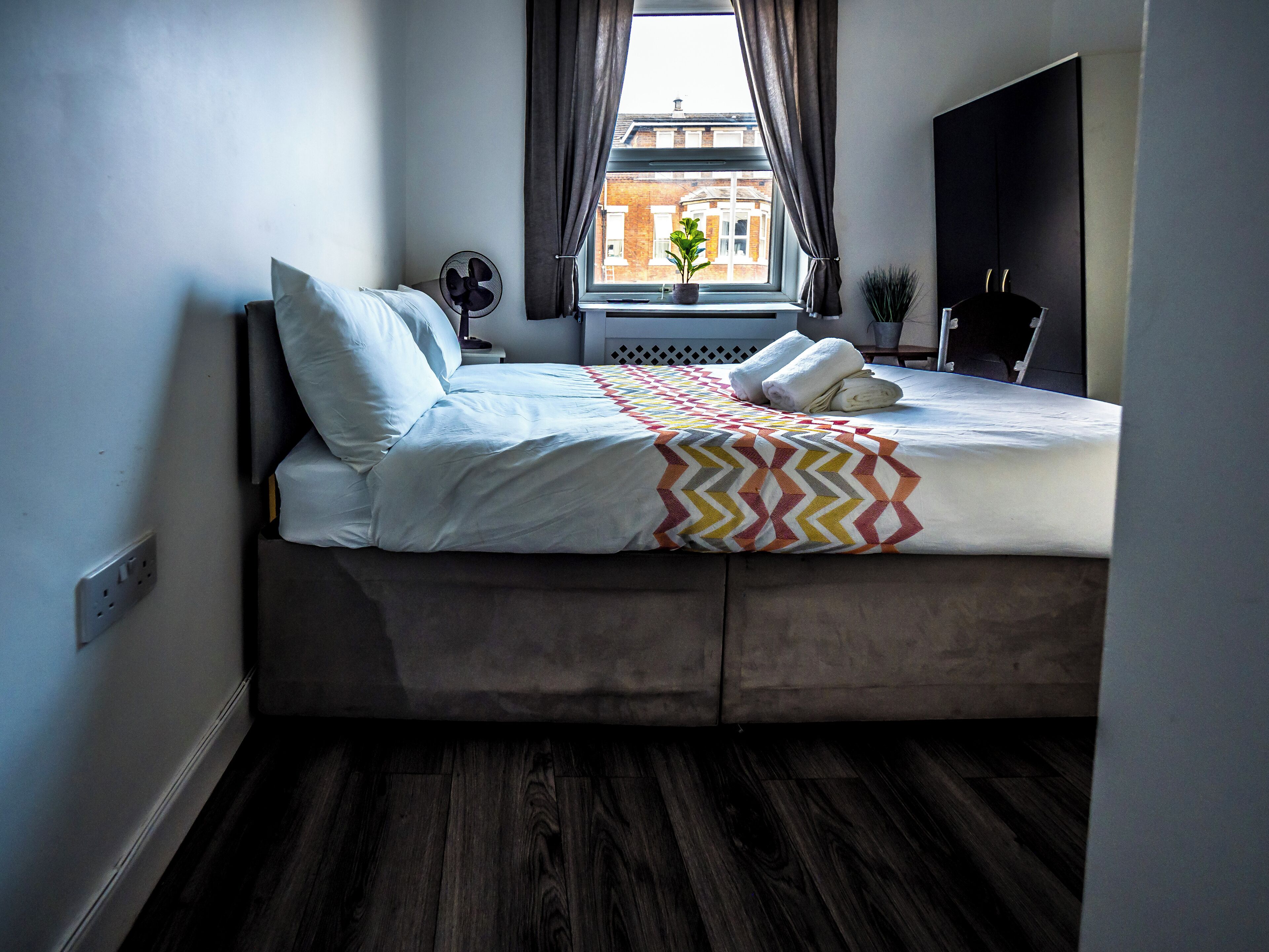 A bedroom with a bed, a window, a plant, and a fan.