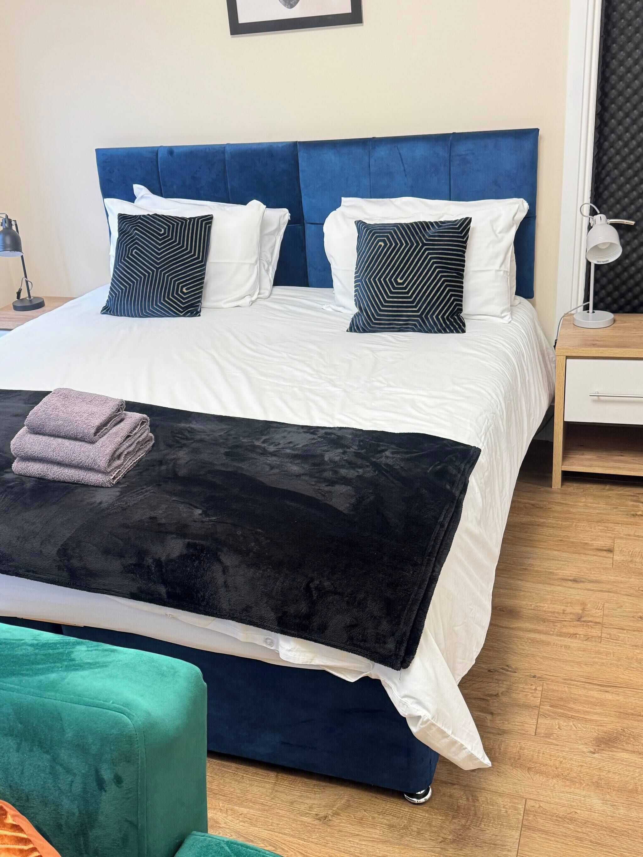 Een net opgemaakt bed met een blauw hoofdeinde, witte beddengoed en gestreepte kussens. Een opgevouwen handdoek ligt op het bed, en een groene fauteuil is op de voorgrond te zien.
