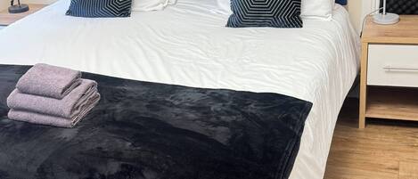 Een net opgemaakt bed met een blauw hoofdeinde, witte beddengoed en gestreepte kussens. Een opgevouwen handdoek ligt op het bed, en een groene fauteuil is op de voorgrond te zien.