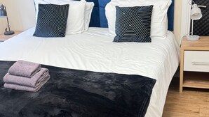 A neatly made bed with a blue headboard, white bedding, and patterned pillows. A folded towel is on the bed, and a green chair is visible in the foreground. - Apartment 2- 216 High Street (Arbroath)