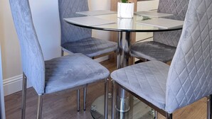 A dining area with a round glass table, four grey upholstered chairs, a white orchid in a marble pot, a flat-screen TV on the wall, and a small potted plant. - Apartment 2- 216 High Street (Arbroath)