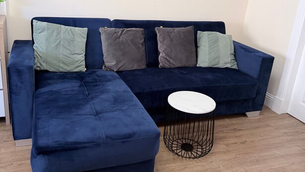 A modern living room with a dark blue sectional sofa, two green and two gray pillows, and a small round coffee table with a white top and black wireframe base. - Apartment 2- 216 High Street (Arbroath)