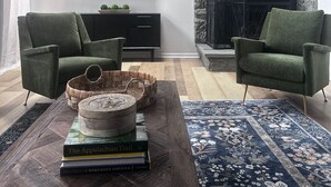 A modern living room with a stone fireplace, two green armchairs, a wooden coffee table with books, and a flat-screen TV mounted on the wall.