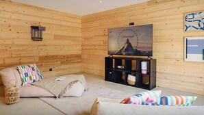A cozy room with wooden panel walls, a flat-screen TV mounted on a black stand, a cushioned seating area with striped pillows, and a woven basket. - Chalet Sheehy - Modern property with spa and terraces - OVO Network (La Toussuire)