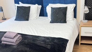 A neatly made bed with a blue headboard, white bedding, and patterned pillows. A folded towel is on the bed, and a green armchair is visible in the foreground.
