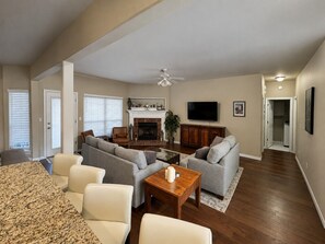 A spacious living room with a fireplace, a television, and a dining area with a granite table. - La Fleur Jenks | 5BR • Near Tulsa & Tanger Outlets (Jenks)