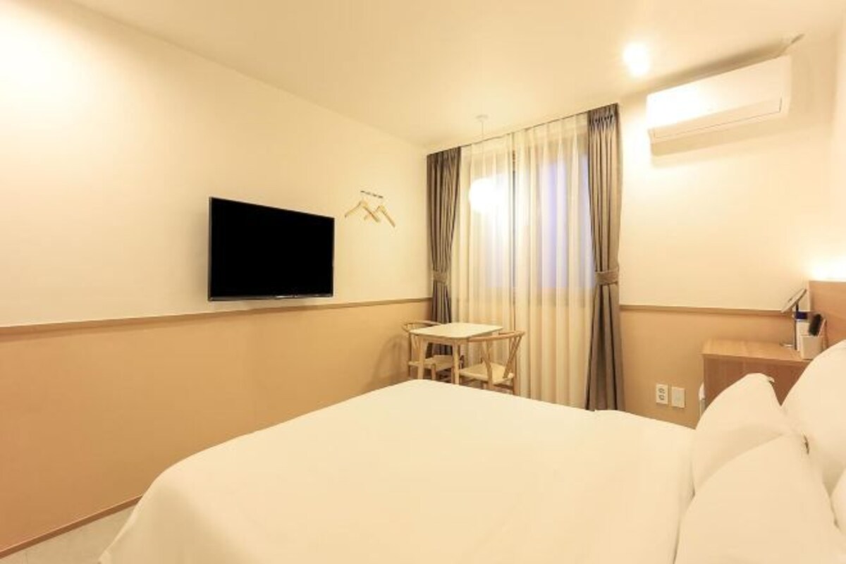 A hotel room with a bed, a TV mounted on the wall, a small table with chairs, and a window with curtains.