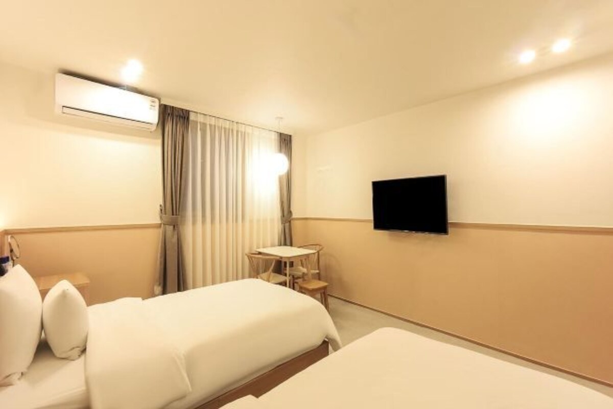 A hotel room with two beds, a TV mounted on the wall, an air conditioning unit, a small table, and a window with curtains.