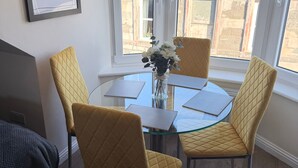 A dining area with a glass table and yellow chairs, a vase with flowers, and a view of buildings through large windows. - Apartment 6- 216 High Street (Arbroath)