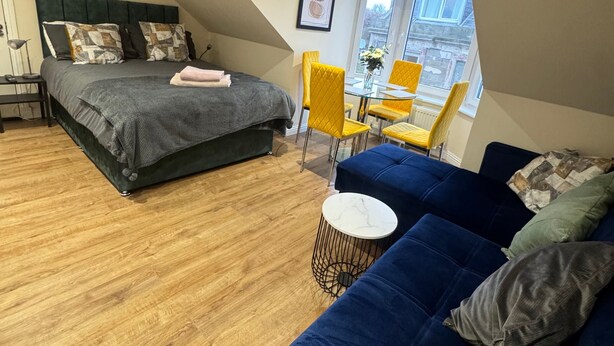 A modern bedroom with a large bed, a blue sofa, and a small table. - Apartment 6- 216 High Street (Arbroath)