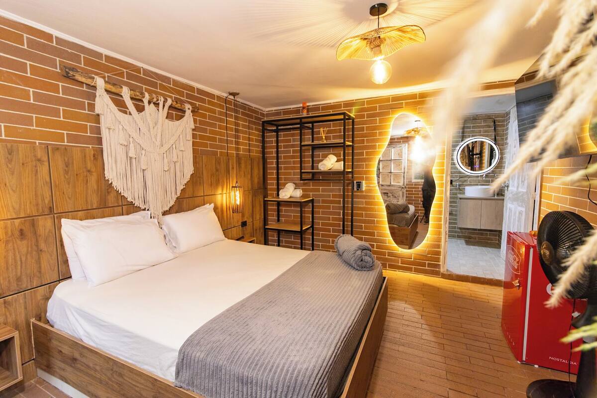 Een slaapkamer met een houten bed, een macramé wanddecoratie, een plank met handdoeken, een spiegel en een rode apparate.