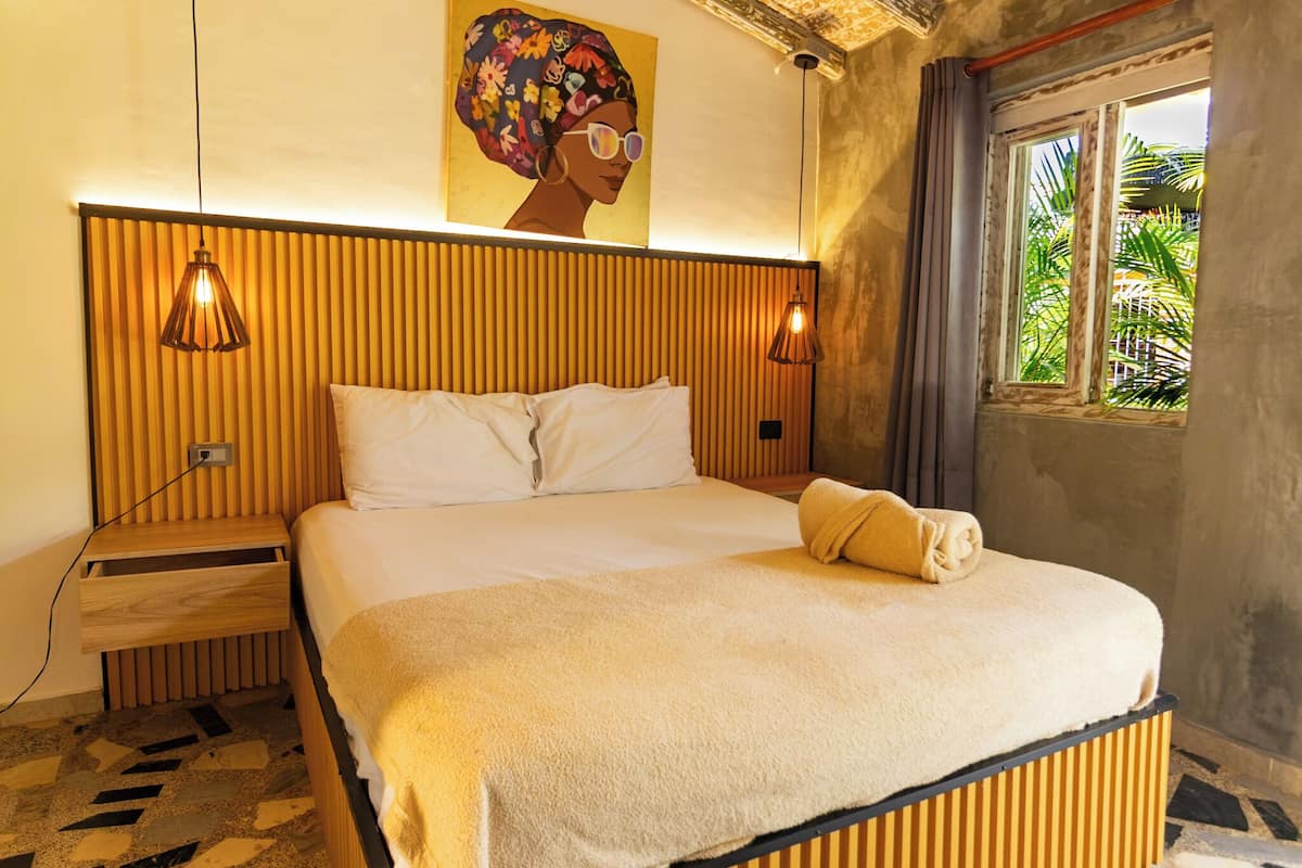 Een hotelkamer met een bed, een houten hoofdeinde, twee bedlampjes, een raam met uitzicht op palmbomen en een schilderij aan de muur.