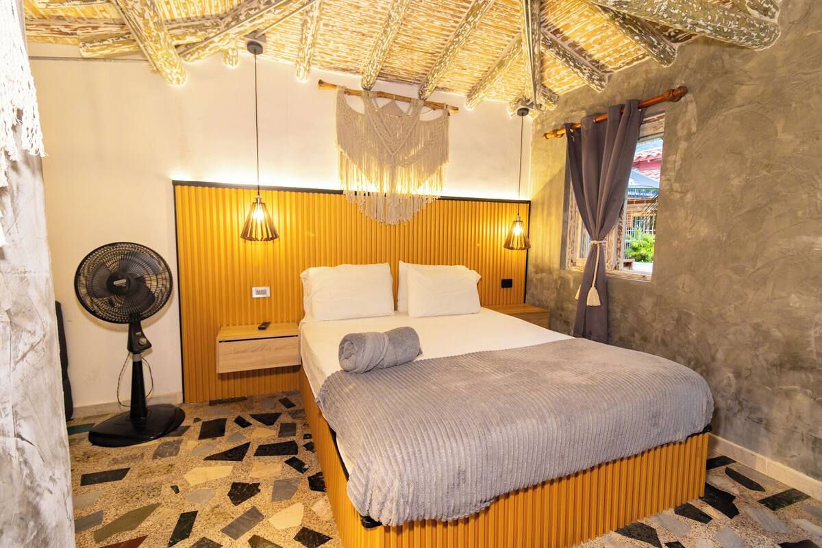 Een kamer met een bed, nachtkastje, ventilator en een raam met gordijnen.