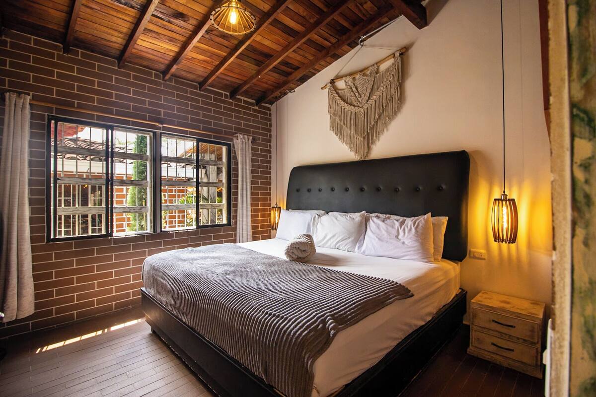 Een slaapkamer met een groot bed, houten balken in het plafond, een bakstenen muur en een raam met gordijnen.