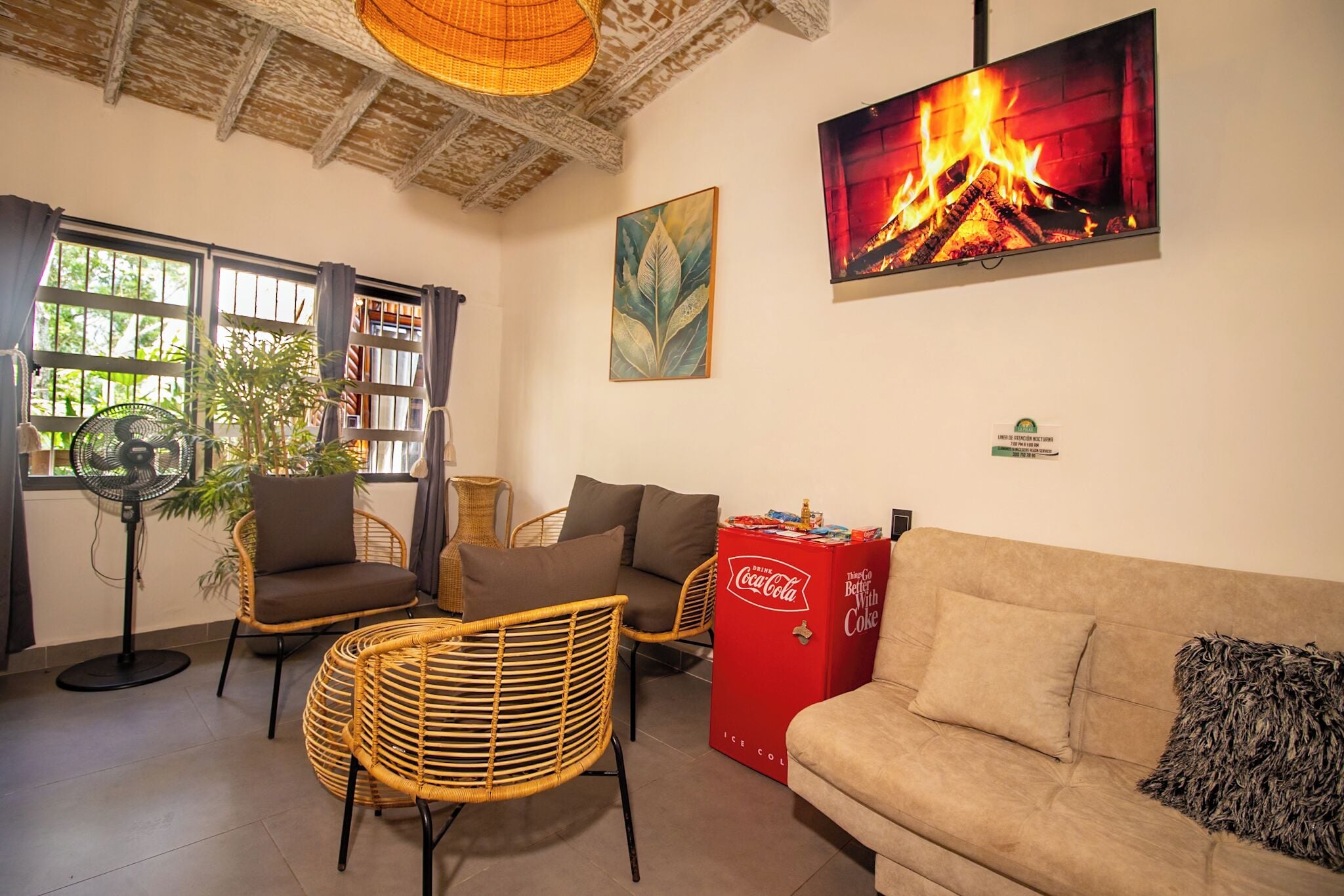 A cozy living area with wicker furniture, a sofa, a fan, a Coca-Cola refrigerator, and a fireplace image on the wall.