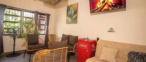 A cozy living area with wicker furniture, a sofa, a fan, a Coca-Cola refrigerator, and a fireplace image on the wall.