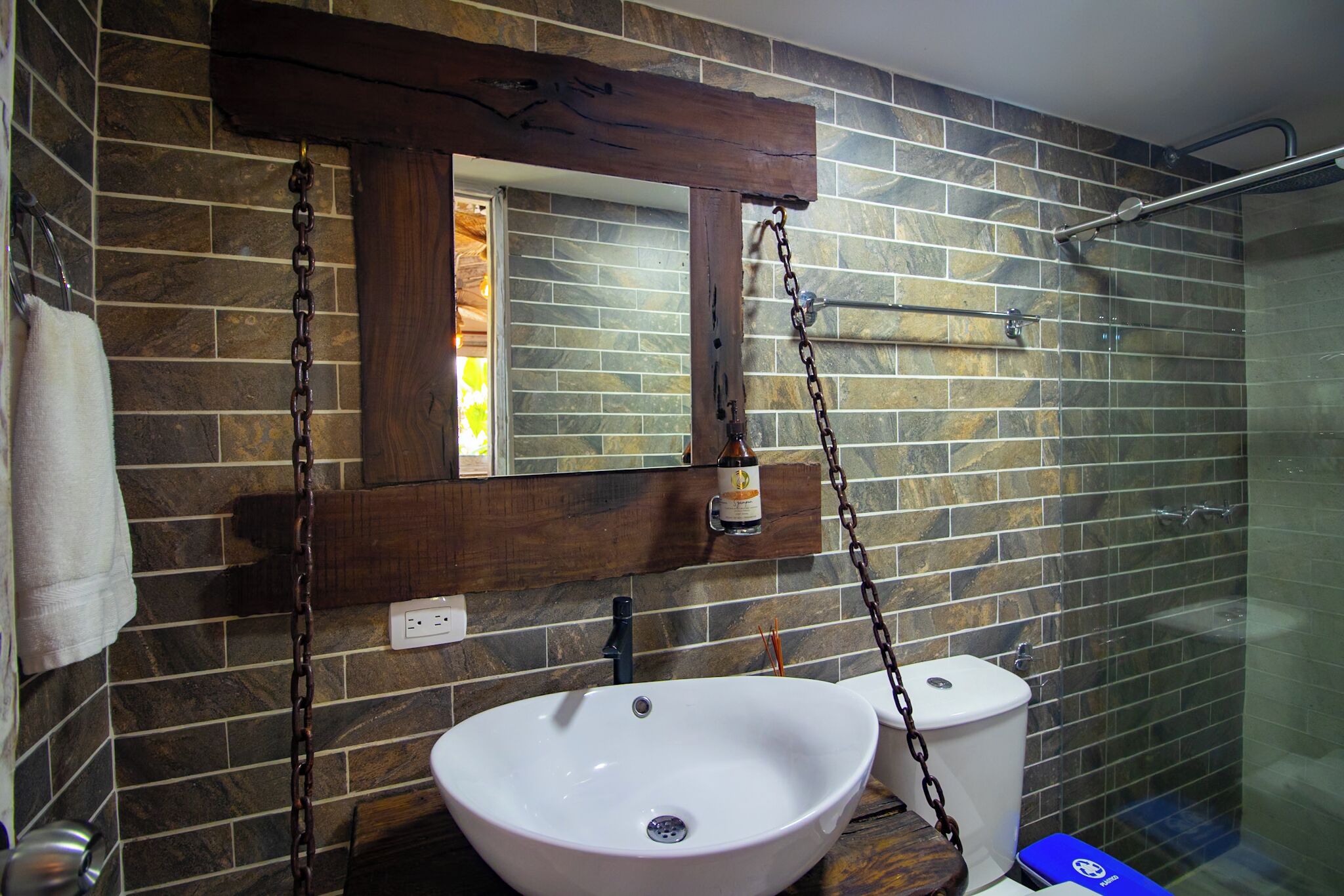 Un baño con pared de baldosas de piedra, un lavabo blanco, un espejo con marco de madera y una cabina de ducha de vidrio.