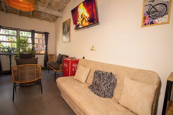 Una sala de estar acogedora con un sofá color beige, un sillón de mimbre, un refrigerador rojo de Coca-Cola y un ventanal con cortinas.