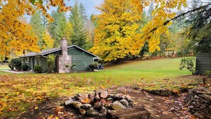 Un chalet rustique avec une cheminée en pierre, entouré d’une cour parsemée de feuilles d’automne et d’une pile de bois de chauffage.