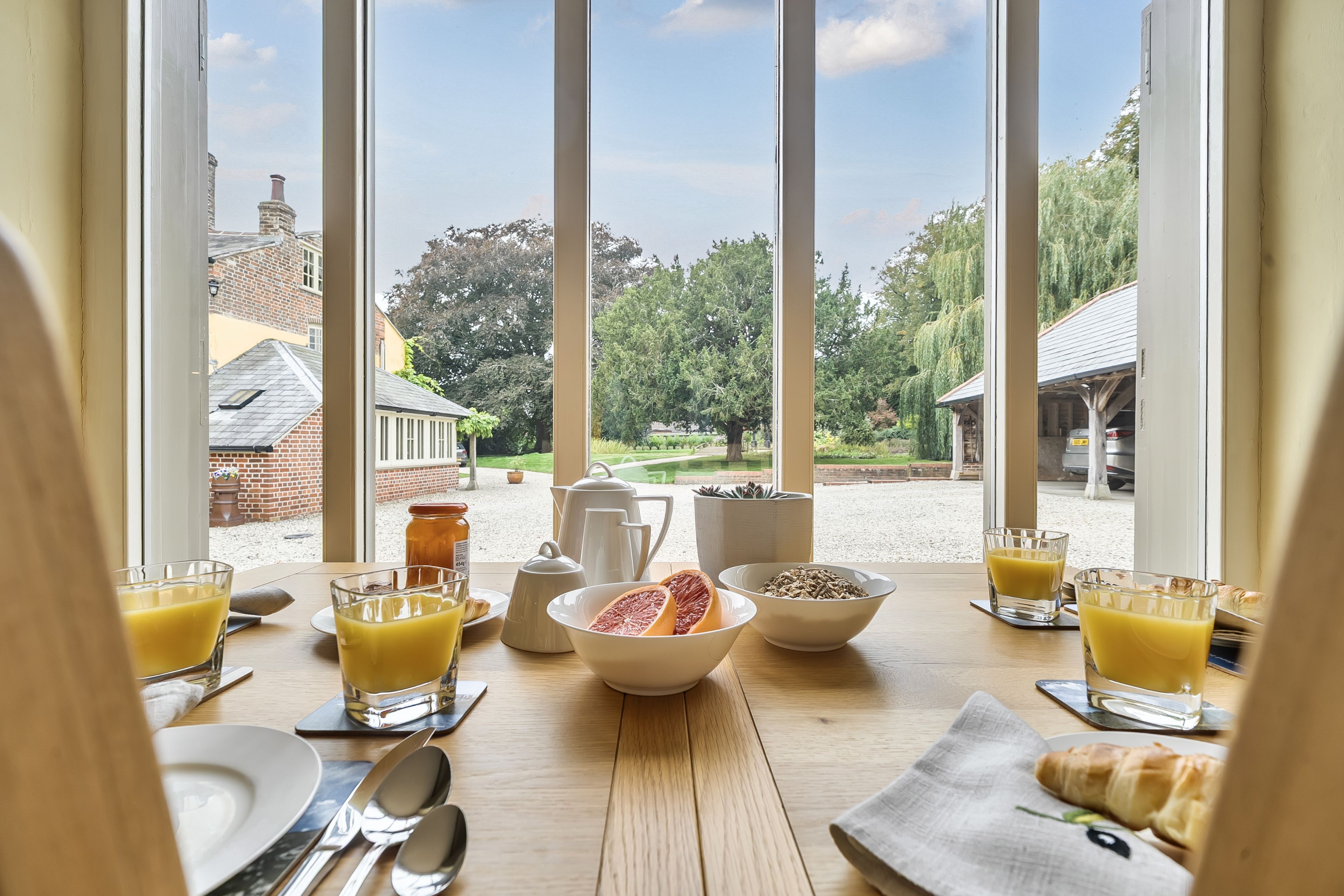 A breakfast table with orange juice, grapefruit, and a croissant, overlooking a garden and buildings.