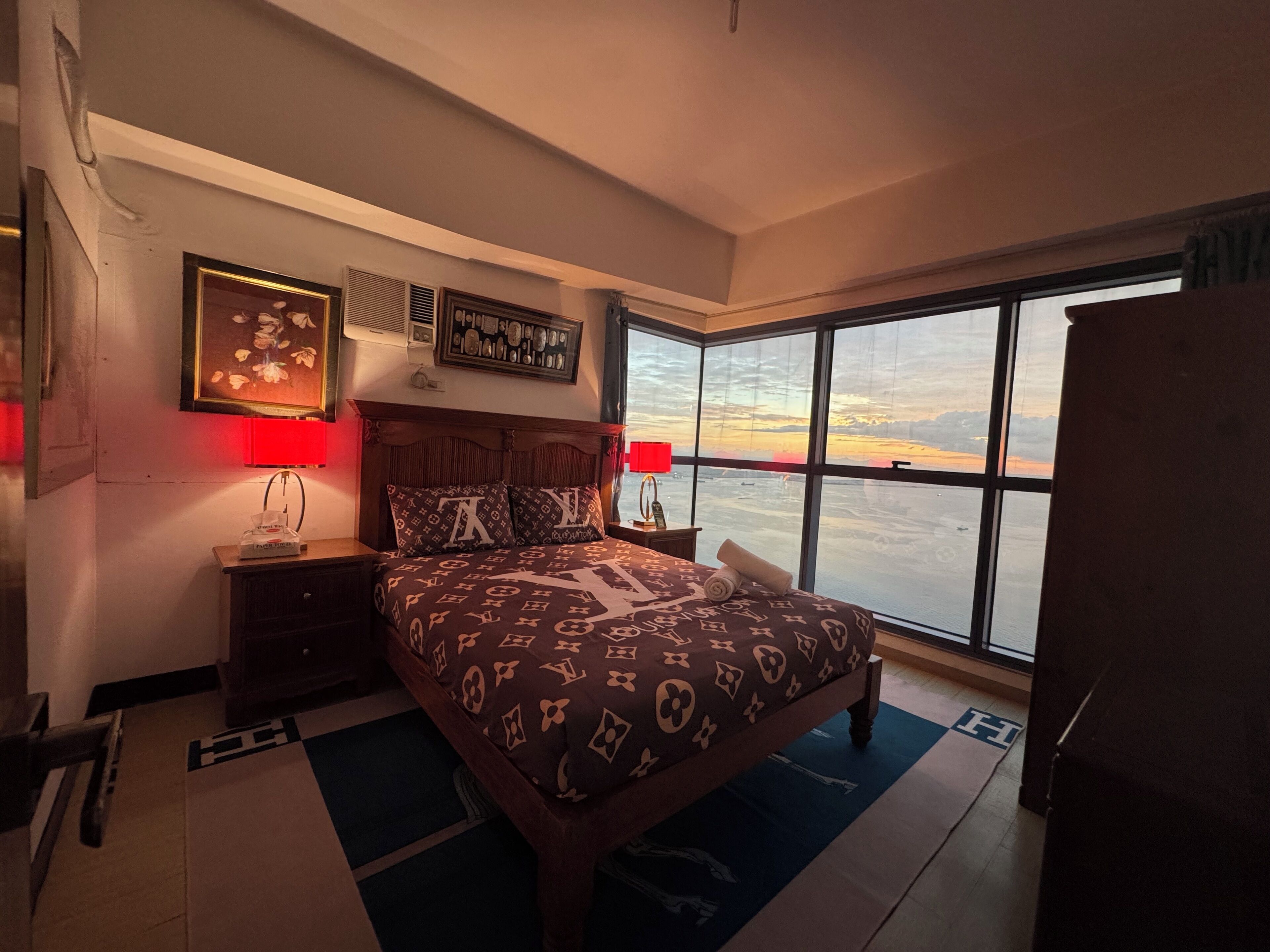 Une chambre avec un grand lit, deux lampes de chevet, une table de chevet, un tableau encadré et une vue sur un coucher de soleil se reflétant sur une étendue d’eau.