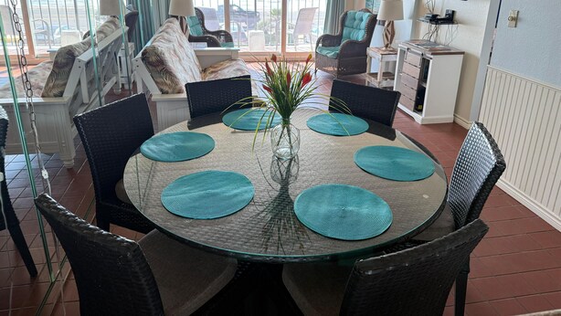A dining area with a glass table, teal placemats, wicker chairs, and a view of the ocean. - Relaxing Beachfront Oceanview Condo w/2 Beds 2 Full Baths, Kitchen, Pet Friendly (Galveston)