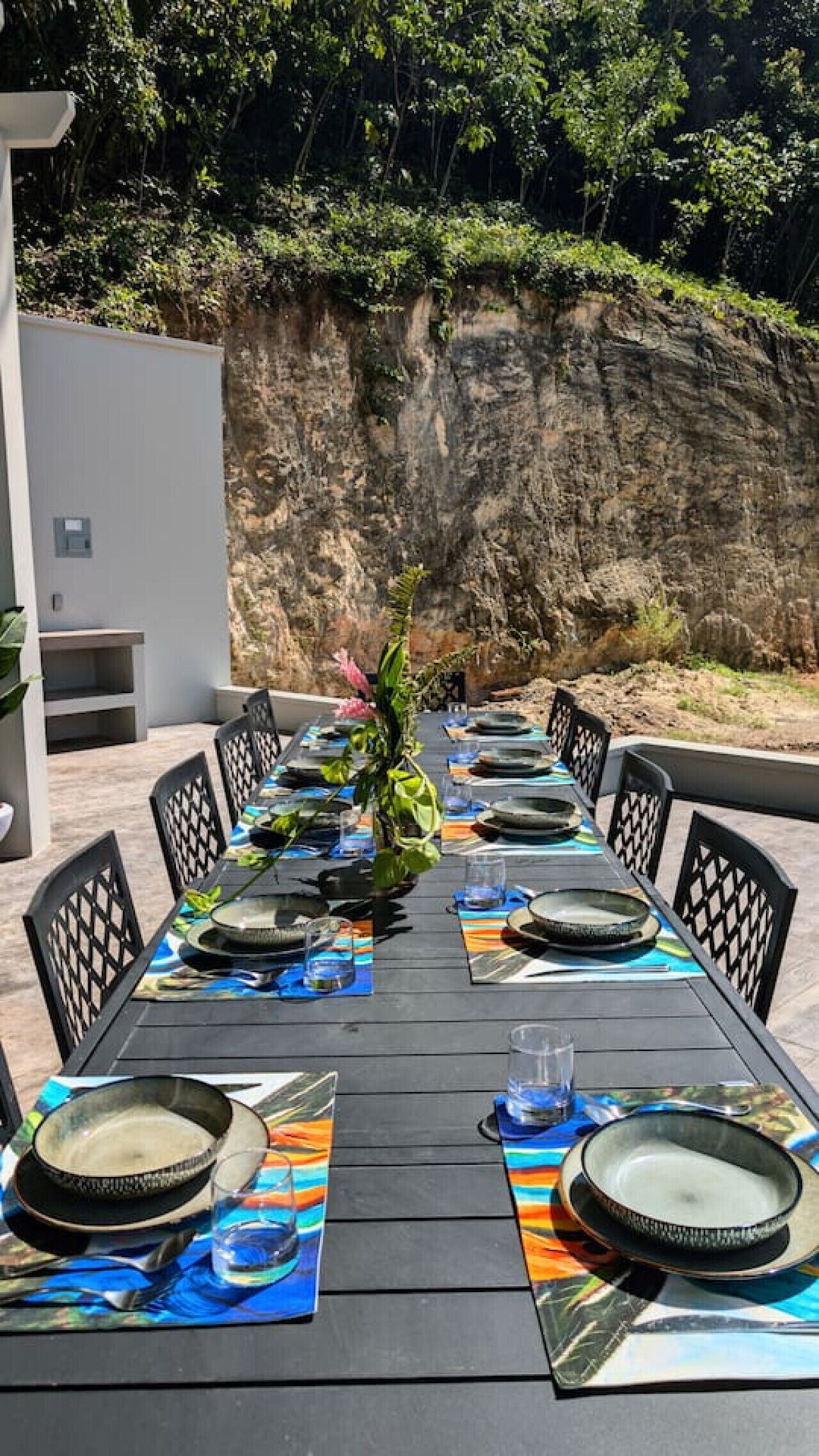An outdoor dining area with a long table set for six, surrounded by chairs, with a rocky cliff in the background.