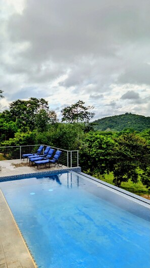 Une piscine bordée de chaises longues bleues, avec vue sur une colline boisée.