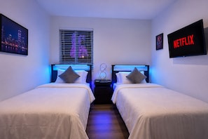 Ein Hotelzimmer mit zwei Betten, einem Fenster mit Jalousien, einem an der Wand montierten Fernseher und einem gerahmten Bild.