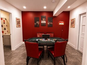 Une pièce avec une table de casino recouverte de feutre vert, des chaises rouges et des œuvres d’art murales comprenant des jetons de poker et un panneau néon.