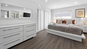 Un dormitorio moderno con una cama grande, muebles blancos, un televisor y una ventana con persianas.