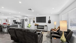 Una sala de estar moderna con un sofá, una mesa de centro y un televisor. Se observa una cocina al fondo con un refrigerador, microondas y estufa.