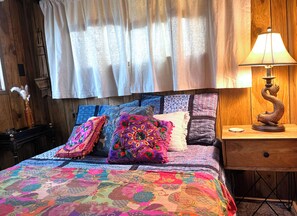 A bed with colorful quilts and pillows, a wooden headboard, a nightstand with a lamp, and a window with curtains. - Cozy Cabin Retreat in the Woods (Edwardsville)