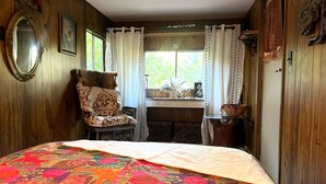 A room with wooden walls, a bed with a colorful quilt, a chair, a desk with a lamp, and a window with curtains.