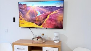 Ein modernes Wohnzimmer mit einem Flachbildfernseher an der Wand, einer Holzkombination mit Schubladen, einer Kaffeemaschine und einem weißen Sessel.