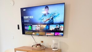 Ein modernes Wohnzimmer mit einem wandmontierten Flachbildfernseher, der eine Auswahl an Filmen zeigt. Ein Holzschrank mit Schubladen und eine Kaffeemaschine stehen darauf. Zwei weiße Sessel befinden sich davor.