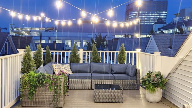 A rooftop deck with a gray sofa, two wicker side tables, and string lights.