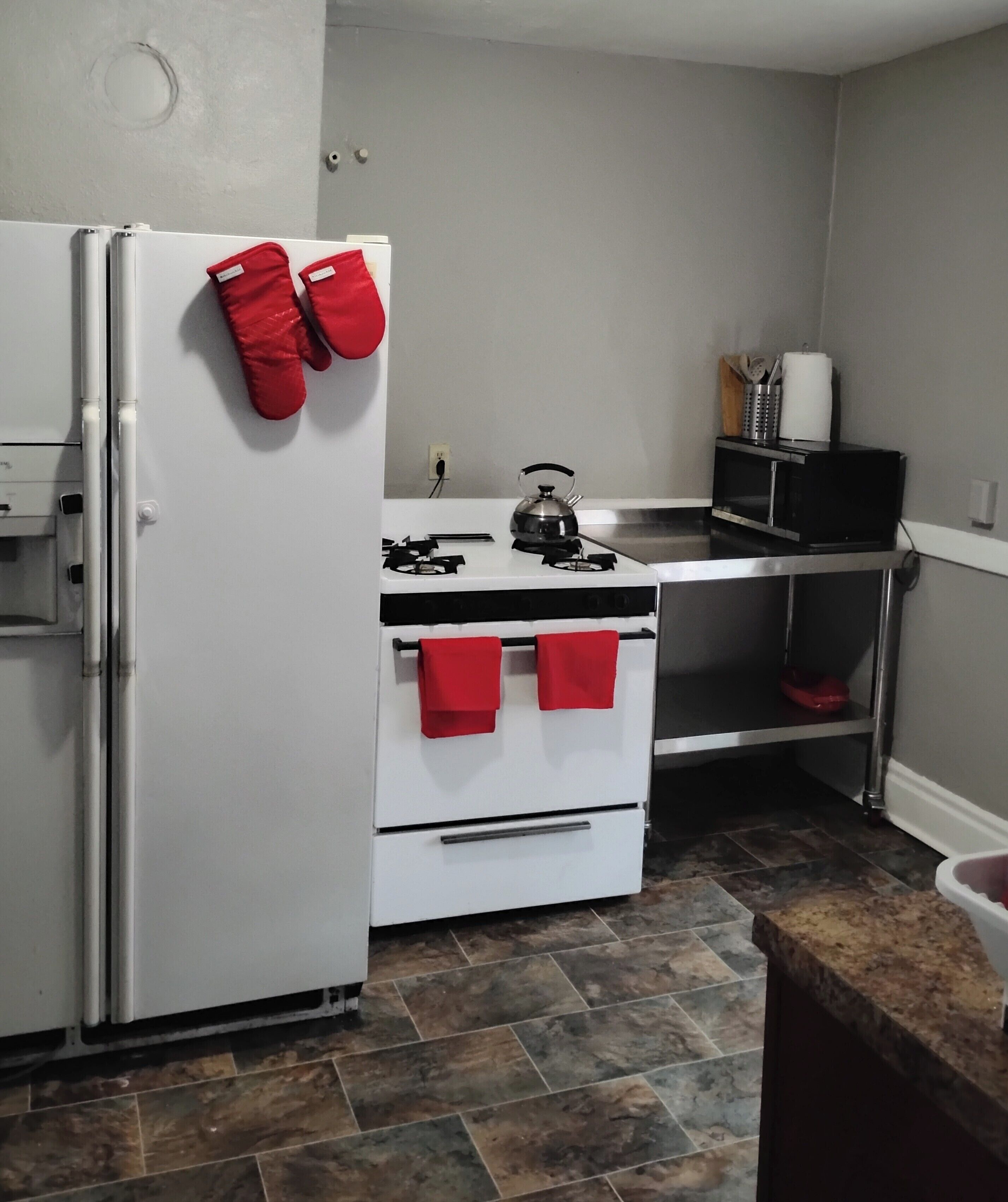 A compact kitchen with a white refrigerator, a stove, and a microwave. Red oven mitts hang on the refrigerator, and a kettle is on the stove.