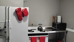 Eine kompakte Küche mit einem weißen Kühlschrank, einem Herd und einer Mikrowelle. Rote Topflappen hängen am Kühlschrank, und ein Wasserkocher steht auf dem Herd.