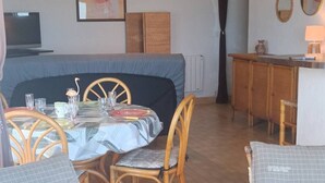 Un comedor con una mesa dispuesta para cuatro personas, rodeada de sillas de mimbre. En primer plano, un sofá con cojines a cuadros. Un televisor está montado en la pared a la izquierda.