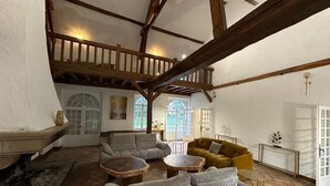 Un vaste espace de vie doté d’un haut plafond, de poutres apparentes et d’une cheminée. On y trouve deux canapés et une table basse, et de grandes fenêtres offrent une vue sur un jardin.