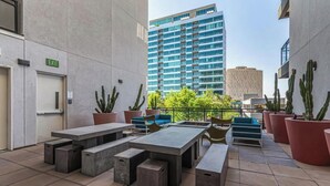 Eine Terrasse im Freien mit Betontischen und -bänken, umgeben von Kakteen in Töpfen, und ein Blick auf ein modernes Gebäude im Hintergrund.