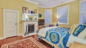 A bedroom with a fireplace, a bed with patterned bedding, a television, and a ceiling fan. - Historic Downtown Victorian Home Near Forsyth (Savannah)