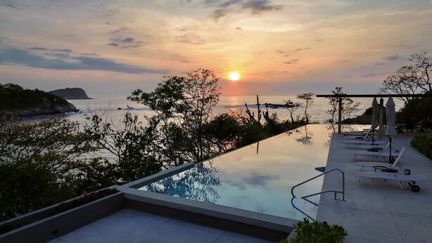 Ein Pool mit Blick auf den Sonnenuntergang über dem Meer, Liegestühle und eine Pergola mit Sonnenschirmen.