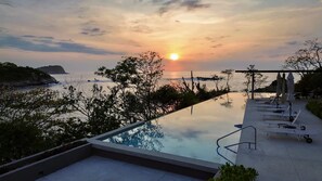 A pool with a view of the sunset over the sea, lounge chairs, and a pergola with umbrellas. - Beautiful luxury ocean front condo. Watch the sun rise out of the pacific ocean! (Huatulco)