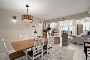 A modern dining area with a wooden table and white chairs, a hanging light fixture, and a view of the living room. - Quay - Luxury Penthouse Condo! (Ocean City)