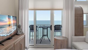Un balcón con vista al mar, un televisor sobre un mueble de madera y dos sillas negras.