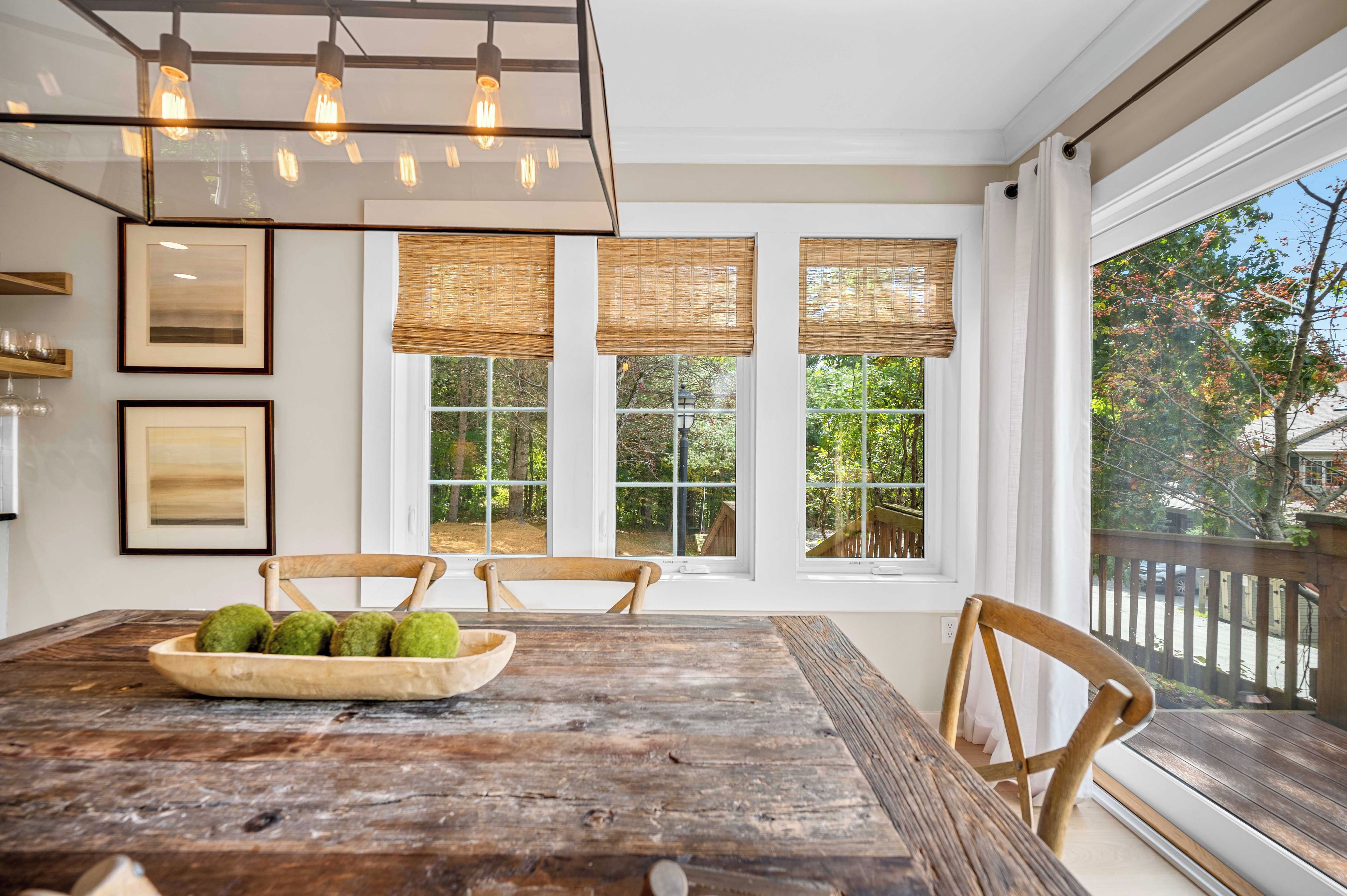 A dining area with a wooden table, chairs, and a bowl of green apples. There are three windows with wicker blinds, and a hanging light fixture above.