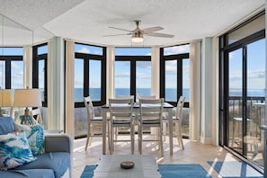 A sunlit room with a dining table and chairs, large windows, and a ceiling fan.